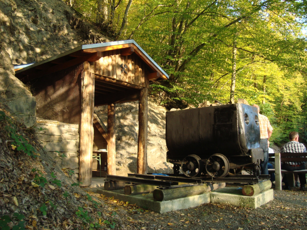 Ein altes aus Holz gebautes Gebäude eines Bergbau Eingangs mit einem Förderwagen der dort als Anschauungsmaterial vor dem Eingang platziert ist.